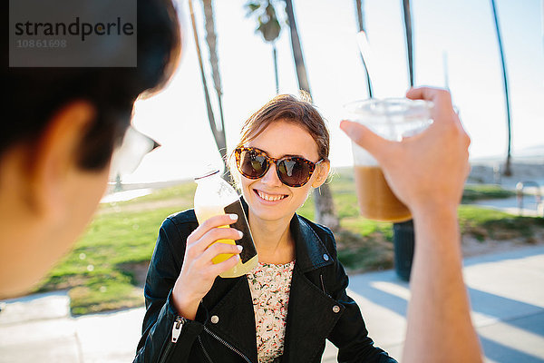 Über-Schulter-Ansicht eines jungen Paares  das einen Saft- und Kaffee-Toast ausbringt  Venice Beach  Kalifornien  USA