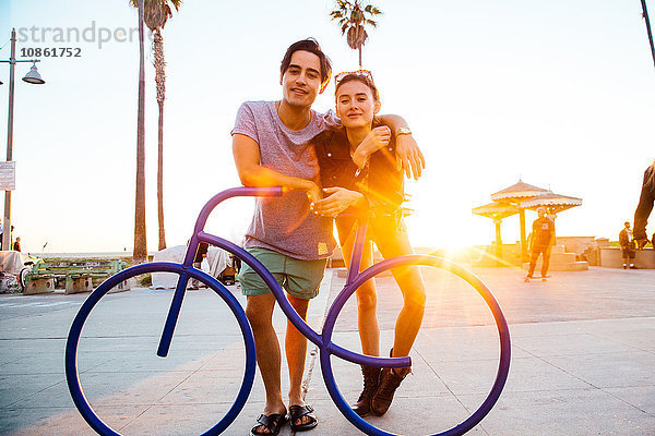 Porträt eines jungen Paares  das sich an eine Fahrradskulptur an der Küste lehnt  Venice Beach  Kalifornien  USA