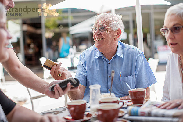 Gruppe reifer Freunde  sitzt vor dem Café  älterer Mann reicht dem Kellner die Kreditkarte