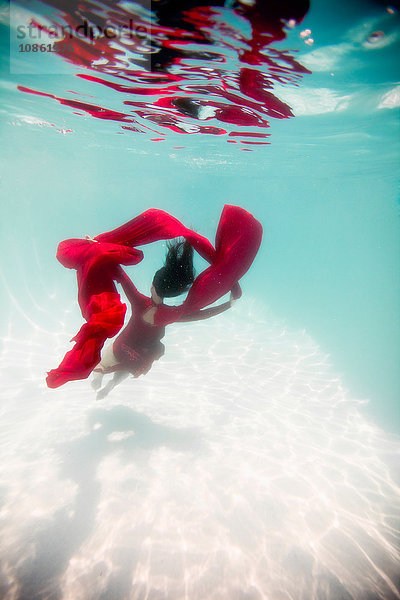 Frau in rotem Kleid  mit rotem Stoff drapiert  schwimmt unter Wasser