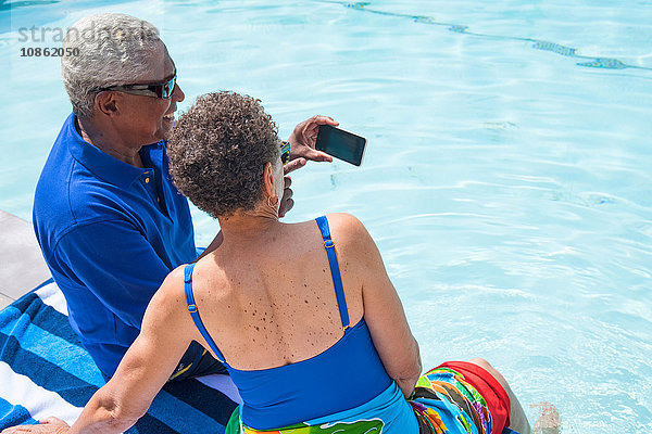 Älteres Ehepaar sitzt neben dem Schwimmbad und benutzt ein Smartphone