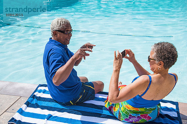 Älteres Ehepaar sitzt am Swimmingpool  Frau fotografiert Mann mit Smartphone