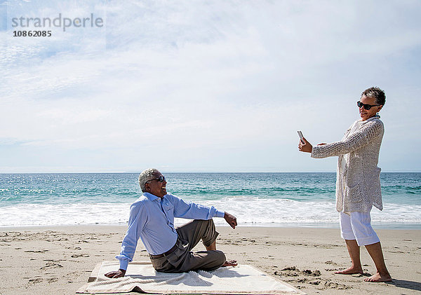 Älteres Ehepaar am Strand  Frau fotografiert Mann mit Smartphone