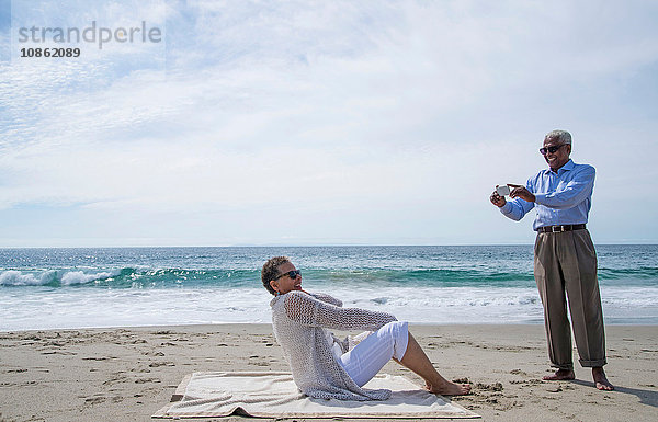 Älteres Ehepaar am Strand  Mann fotografiert Frau mit Smartphone