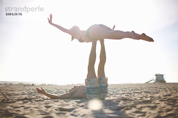Mann liegt am Strand und balanciert Frau auf Füßen