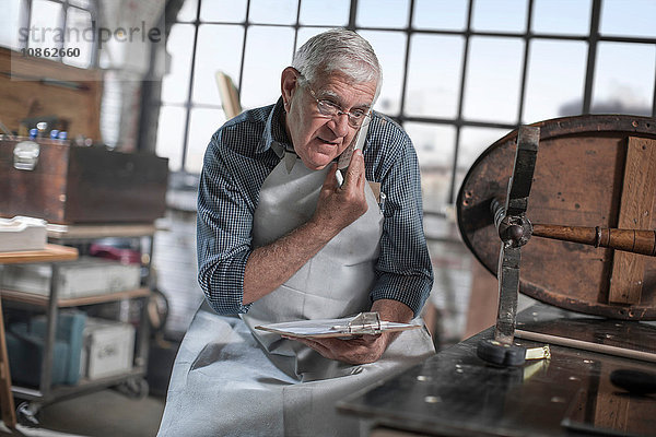 Senior-Handwerker spricht auf Smartphone in einer Antiquitäten-Restaurierungswerkstatt
