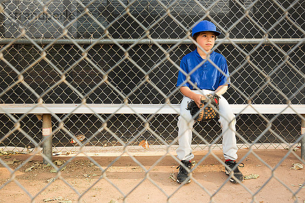 Junge sitzt beim Baseball-Training auf Bank hinter Drahtzaun