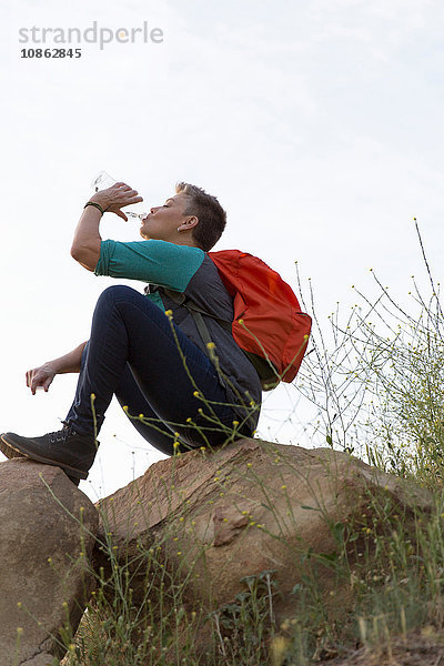 Wanderfrau sitzt auf Felsen und trinkt Wasser aus Flasche