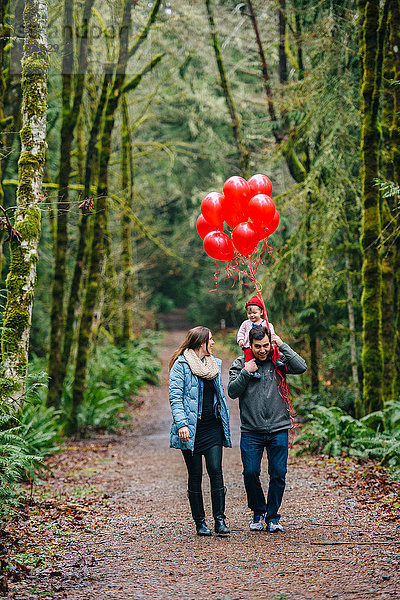 Mittelgroßes erwachsenes Paar trägt Kleinkind-Tochter auf den Schultern mit einem Haufen roter Luftballons im Wald