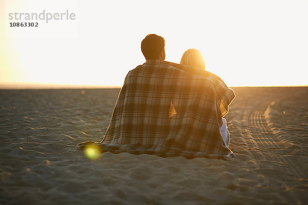 Paar am Strand  in eine Decke gewickelt  Sonnenuntergang beobachtend  Rückansicht