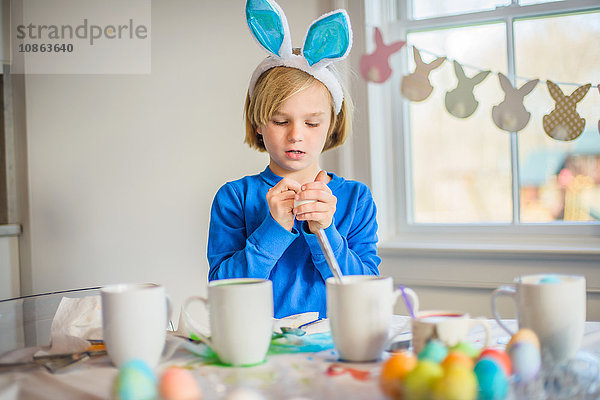 Junge am Tisch mit Hasenohren  die Eier für Ostern schmücken