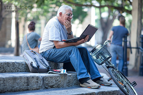 Älterer Mann auf Stadttreppe  der einen Laptop benutzt und verwirrt aussieht