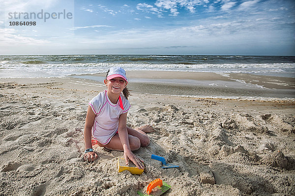 Mädchen sitzt am Strand  spielt im Sand und schaut lächelnd in die Kamera