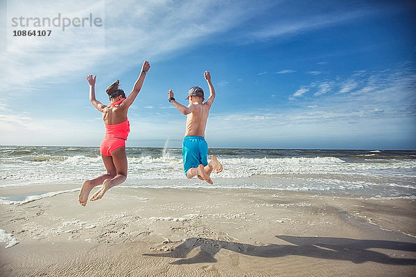 Rückansicht eines Mädchens und eines Jungen am Strand mit erhobenen Armen  die in die Luft springen