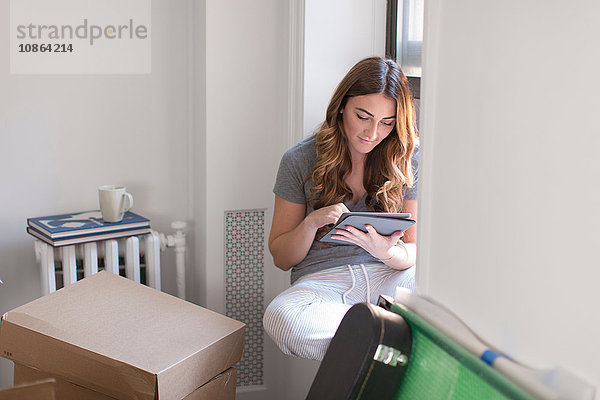 Junge Frau sitzt auf der Fensterbank ihres neuen Zuhauses  benutzt digitales Tablett  Kartons im Zimmer