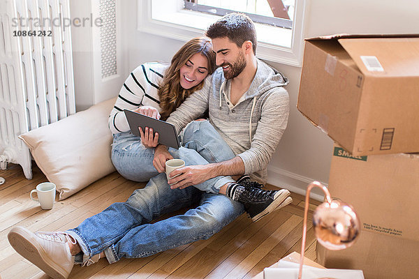 Umzug: Junges Paar sitzt in einem Zimmer voller Kisten und schaut auf ein digitales Tablett