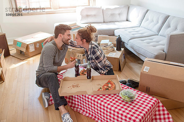 Umzug: Junges Paar isst Pizza und umarmt sich  in neuem Zuhause  umgeben von Kartons