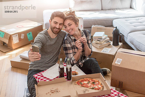 Umzug: Junges Paar beim Selbstporträt mit Smartphone  im neuen Zuhause  umgeben von Kisten