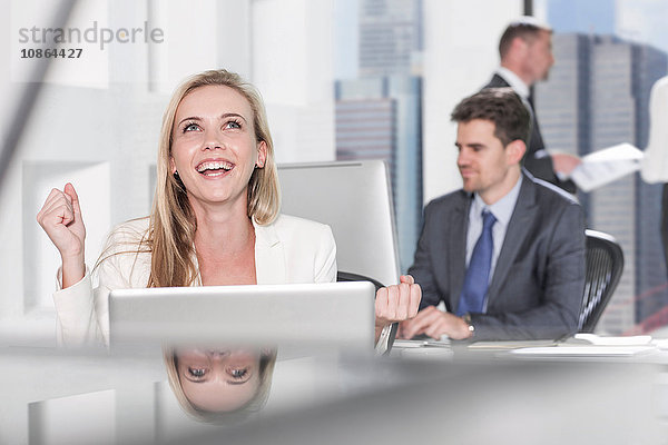 Aufgeregte Geschäftsfrau lächelt breit im Büro  Kollegin arbeitet im Hintergrund