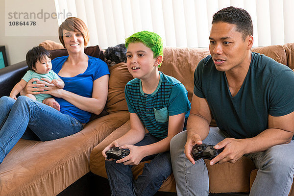Mutter mit Baby beobachtet Vater und Sohn beim Spielen eines Videospiels auf dem Sofa