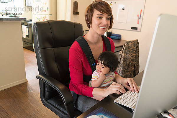 Mutter mit Baby in Tragetasche  am Computer arbeitend
