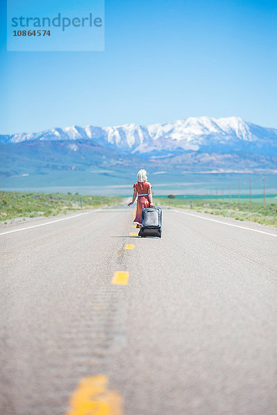 Rückansicht einer jungen Frau im Stil der 1950er Jahre  die allein auf dem Highway 50 geht und einen Rollkoffer zieht  Nevada  USA