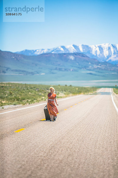 Junge Frau im Stil der 1950er Jahre  die allein auf dem Highway 50 geht und einen Rollkoffer zieht  Nevada  USA