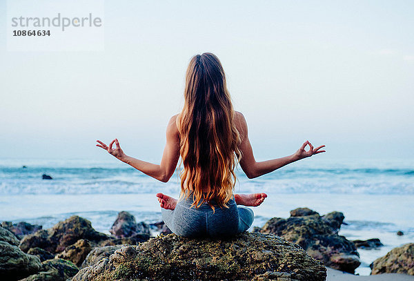 Rückansicht einer jungen Frau mit langen Haaren  die eine Lotus-Yoga-Pose auf Felsen am Strand praktiziert  Los Angeles  Kalifornien  USA