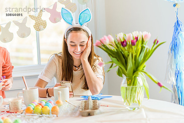 Teenager-Mädchen liest am Tisch Smartphone-Texte und färbt hart gekochte Eier für Ostern