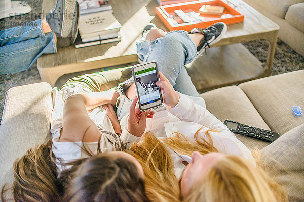 Draufsicht auf Mutter und Teenager-Tochter  die auf dem Sofa sitzen und auf das Neugeborenenfoto auf dem Smartphone schauen