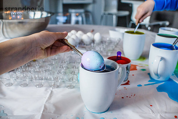 Nahaufnahme der Hand eines Jungen  der zu Ostern ein blau gefärbtes Ei aus einem Becher auf dem Tisch entfernt