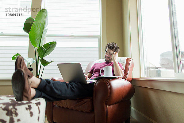 Mann entspannt sich auf Wohnzimmersessel mit den Füßen nach oben und stöbert am Laptop