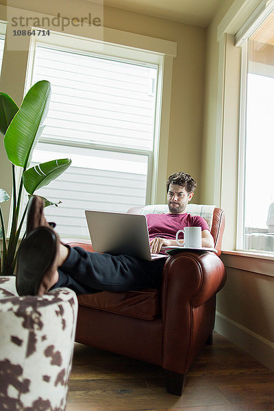 Mann auf Wohnzimmersessel mit den Füßen nach oben beim Arbeiten am Laptop