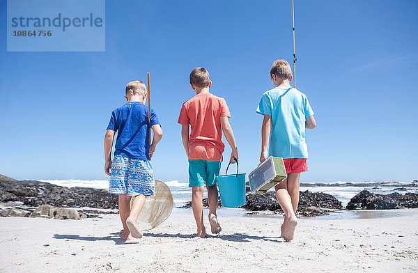 Rückansicht von drei Jungen  die mit Fischernetz  Eimer und Angelrute am Strand spazieren gehen  Kapstadt  Südafrika
