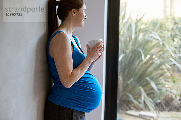 Seitenansicht einer schwangeren Frau  die an der Wand lehnt und eine Kaffeetasse hält und lächelnd aus dem Fenster schaut