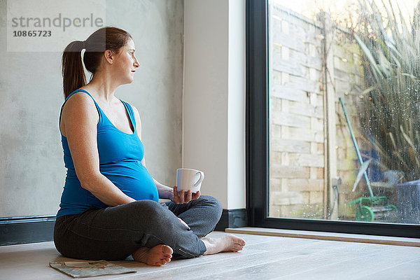 Schwangere Frau sitzt auf dem Boden mit gekreuzten Beinen und hält eine Kaffeetasse und schaut aus dem Fenster