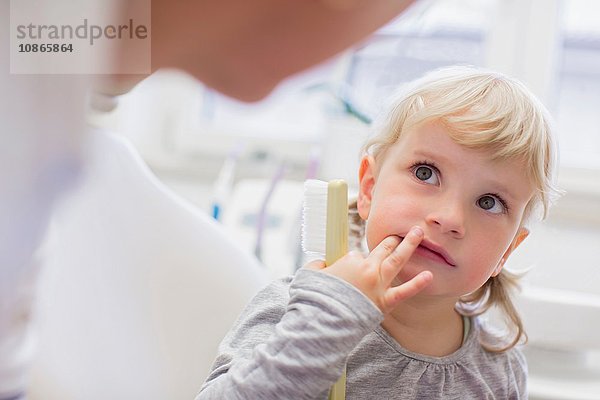 Mädchen mit Fingern im Mund hält Zahnbürste und schaut zum Zahnarzt auf