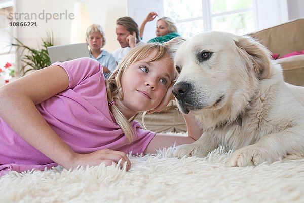 Mädchen und Haushund auf Teppich liegend  Familie mit Laptop im Hintergrund