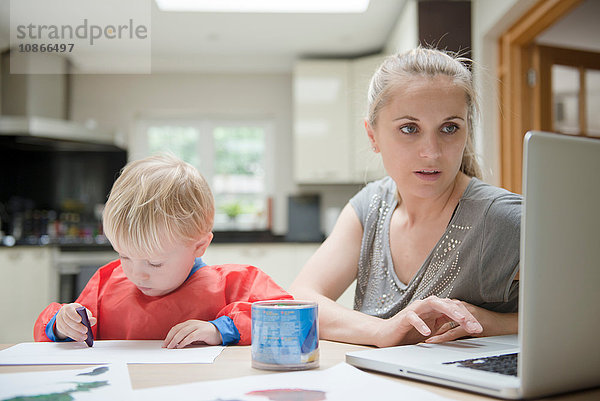 Mutter benutzt Laptop  während der Sohn neben ihr auf Papier zeichnet