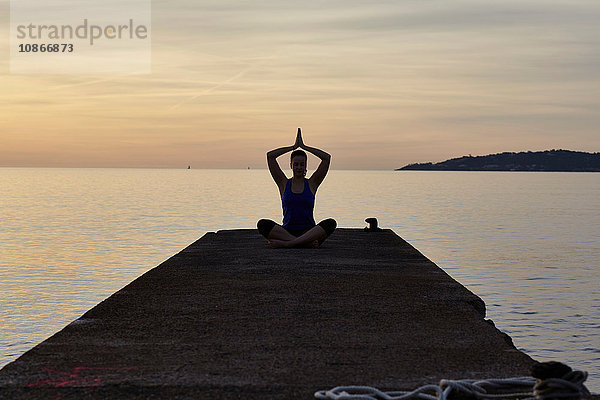 Junge Frau am Pier sitzend  in Yogastellung  in der Abenddämmerung