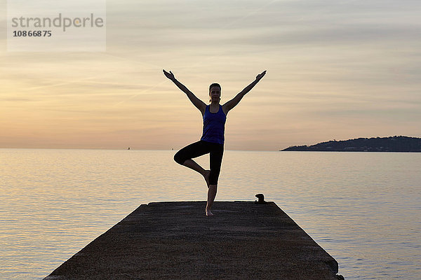 Junge Frau am Pier stehend  in Yogastellung  bei Sonnenuntergang