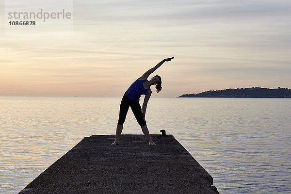 Junge Frau am Pier stehend  in Yogastellung  bei Sonnenuntergang