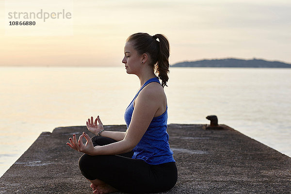 Junge Frau am Pier sitzend  in Yogastellung  bei Sonnenuntergang