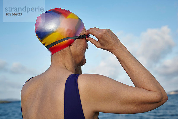 Reife Frau in Badeanzug und Badehut  am Meer stehend  Schwimmbrille anpassen  Rückansicht