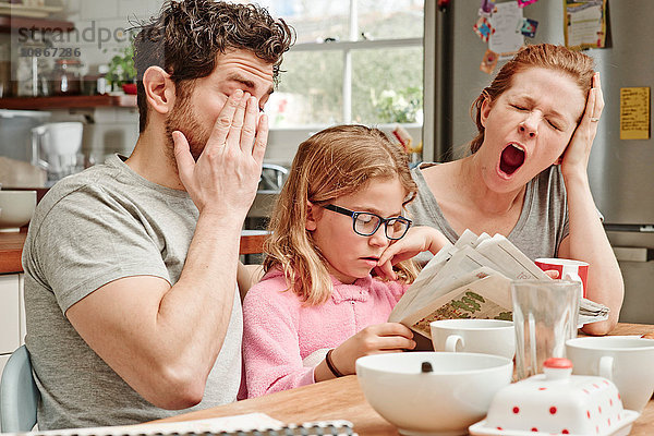 Müde  mittlere erwachsene Eltern am Frühstückstisch  während die Tochter Zeitung liest