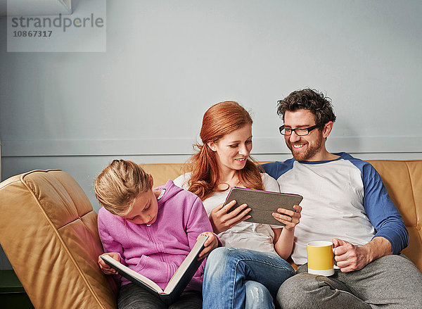 Mittlere erwachsene Eltern und Tochter auf dem Sofa auf Lesebuch und digitalem Tablett
