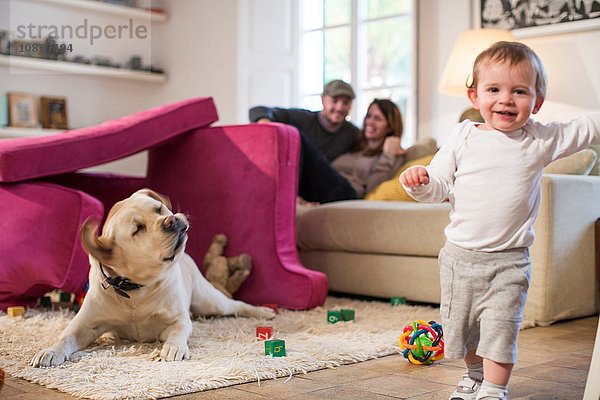 Babyjunge und Haushund spielen in einer Festung aus Sofakissen und schauen lächelnd in die Kamera