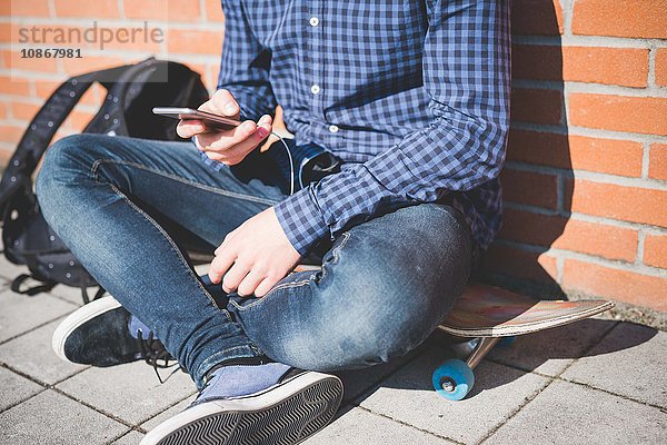 Neck down view of young male urban skateboarder sitting on sidewalk reading smartphone text