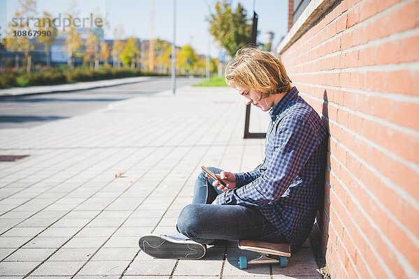 Junge männliche städtische Skateboarder sitzen auf dem Bürgersteig Auswahl Smartphone-Musik