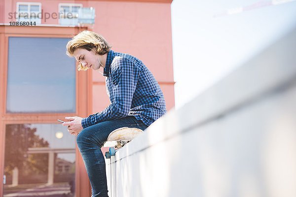 Junge männliche städtische Skateboarder sitzen auf dem Skateboard und lesen Smartphone-Text.